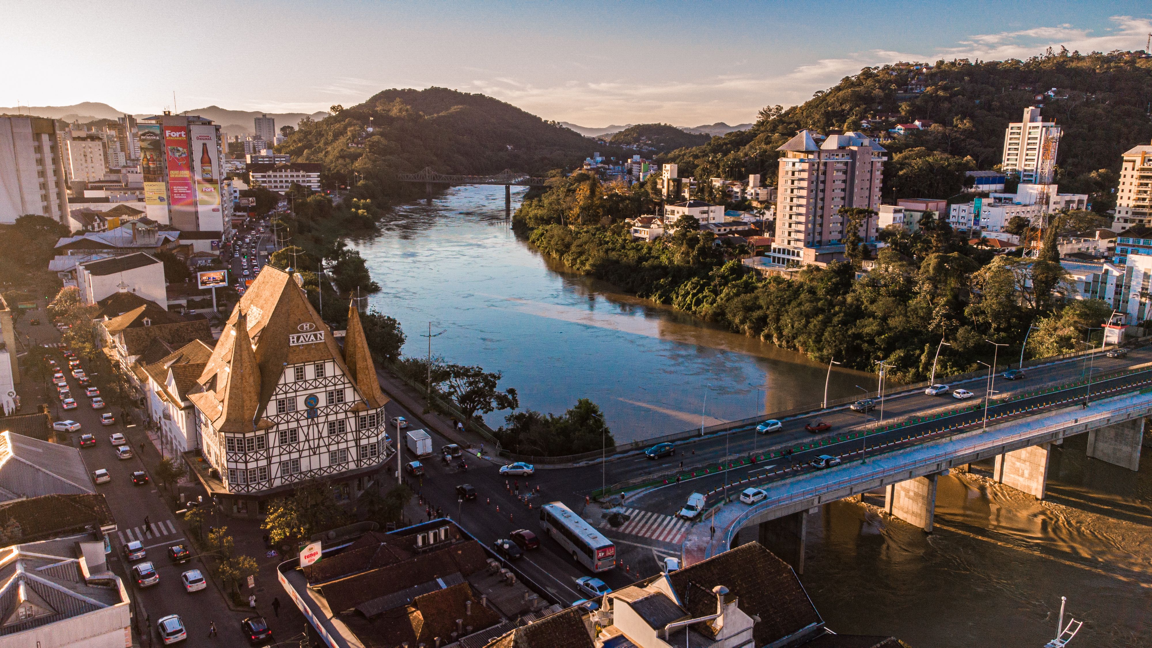 Vista aérea de Blumenau ao entardecer com o Rio Itajaí-Açu, a arquitetura enxaimel e a ponte